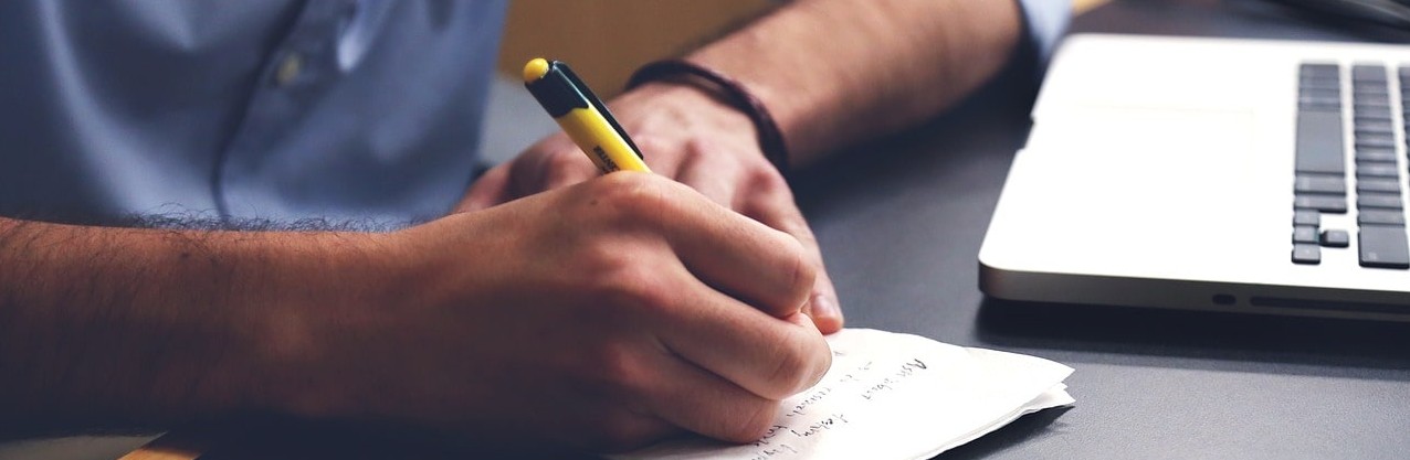 Mann schreibt etwas auf einen Zettel vor seinem Laptop